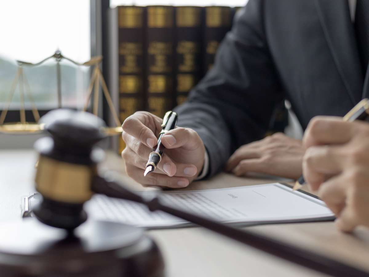 A close-up of a person in a business suit signing a document, with a wooden gavel and the scales of justice in the background, symbolizing the business collections litigation process